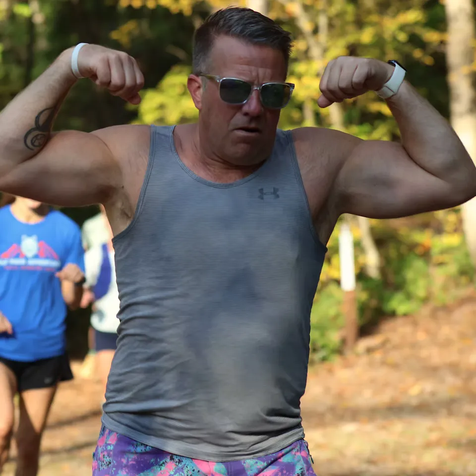 A male runner flexing his biceps while running on a trail, wearing sunglasses and a grey tank top.