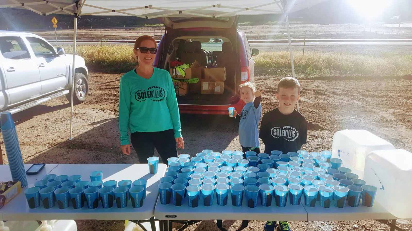 A woman and two children stand behind a table filled with cups at a race aid station.