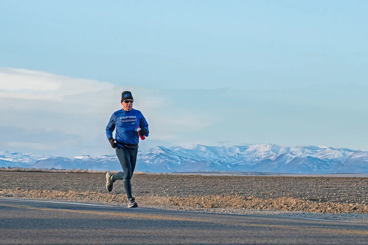 A male runner wearing a blue long-sleeved shirt with 'California Marathon' text and a black beanie runs along a paved road with snow-capped mountains in the background.