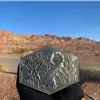 A hexagonal race medal with intricate relief carvings, held up against a backdrop of arid desert mountains and a clear blue sky.