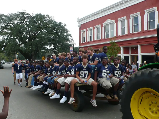 A group of young people in football uniforms sitting on a flatbed trailer pulled by a tractor, with spectators in the background.