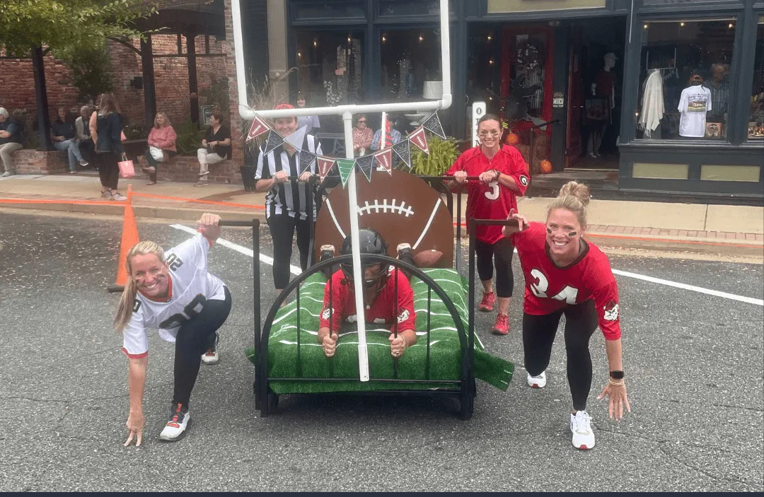People in football jerseys pushing a bed decorated with a football theme, with a goalpost in the background, suggesting a fun, celebratory event.