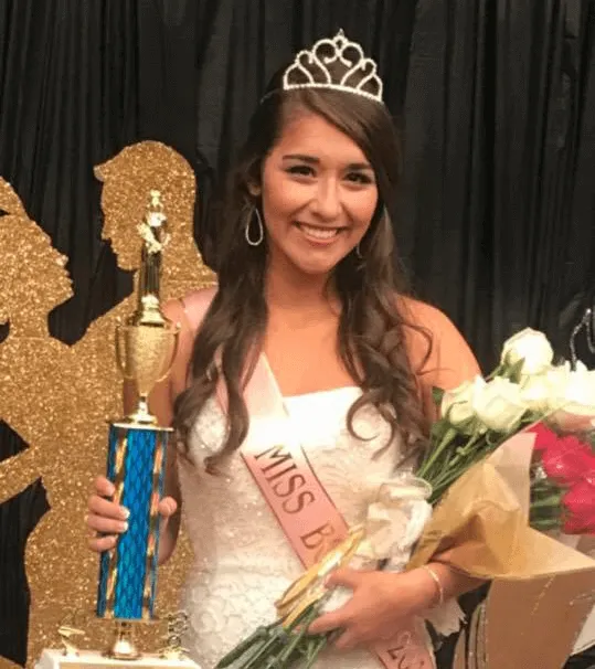A smiling young woman wearing a tiara and a sash that says 'MISS B', holding a trophy and a bouquet of flowers.
