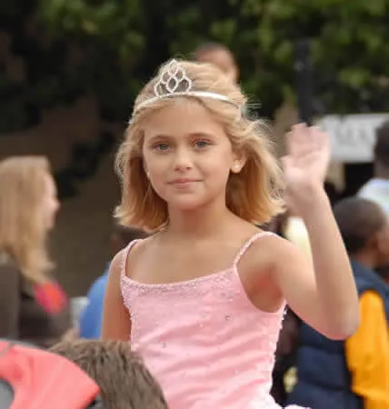 A young girl wearing a tiara and a pink dress waves to the camera.