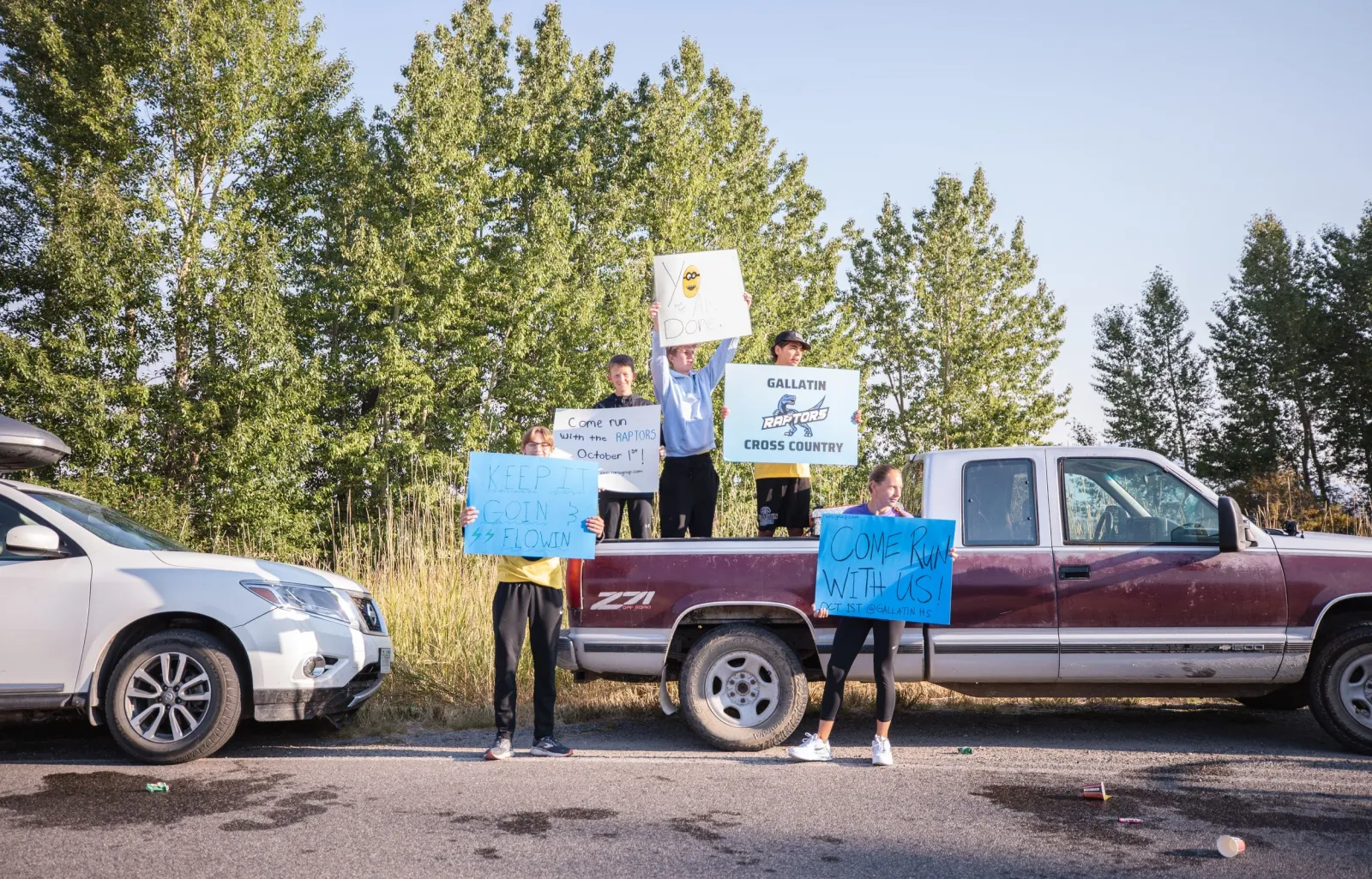 A group of people stand by a pickup truck holding signs that say 'Come Run With Us!' and 'Keep It Goin & Flowin', encouraging participation in the Bozeman Half Marathon and 10K.