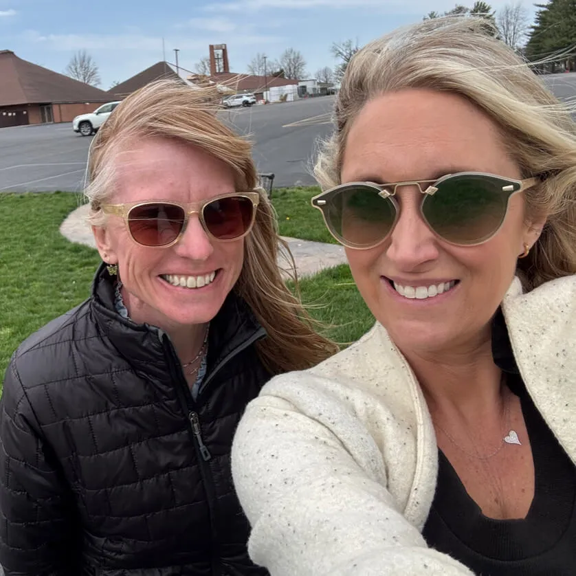 Two smiling women wearing sunglasses pose for a selfie outdoors.