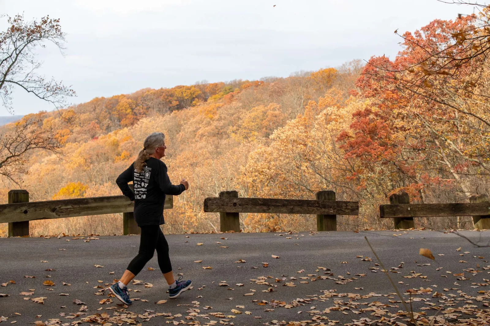 A runner with gray hair tied in a ponytail runs on a paved road with a scenic view of autumn foliage on a hillside.