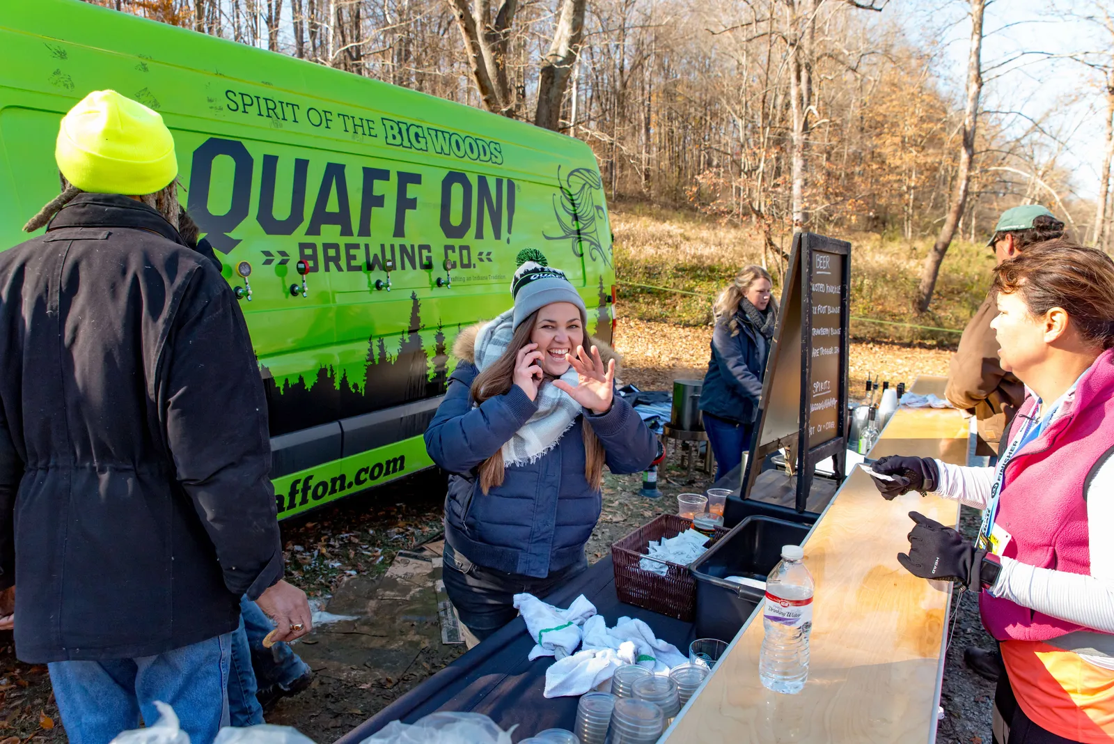 A woman in a blue jacket and grey hat smiles and waves while talking on her phone, standing near a green brewing company van and a refreshment stand at an outdoor event.