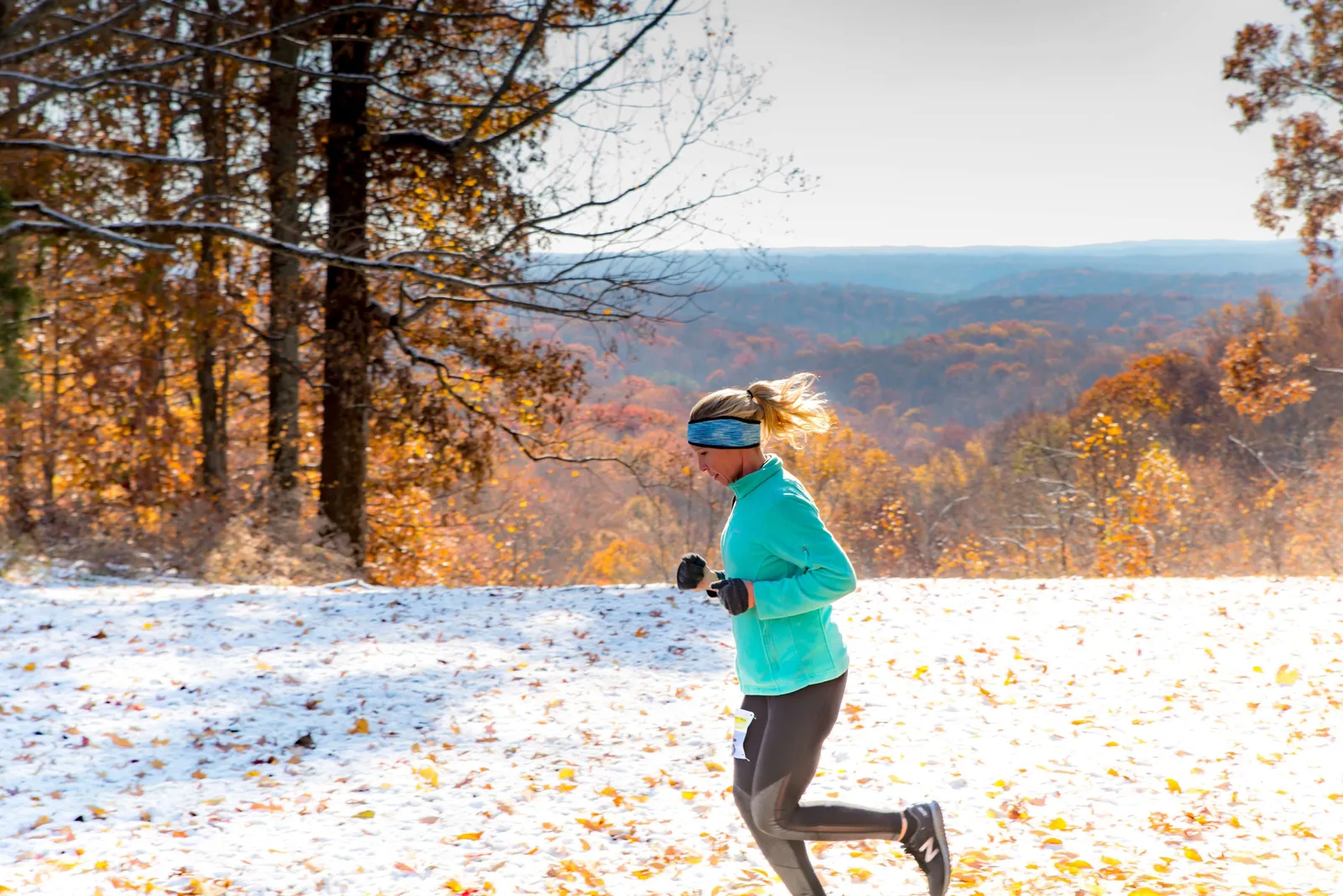 A female runner with blonde hair in a ponytail, wearing a teal jacket and black leggings, runs through a snowy landscape with fall foliage in the background.