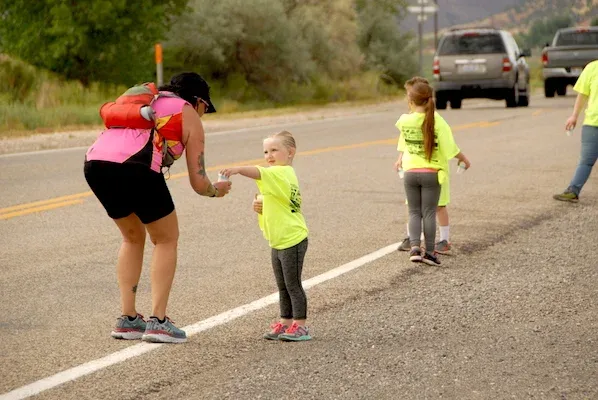 A race volunteer in a pink vest hands a cup to a young runner in a neon yellow shirt on a road.