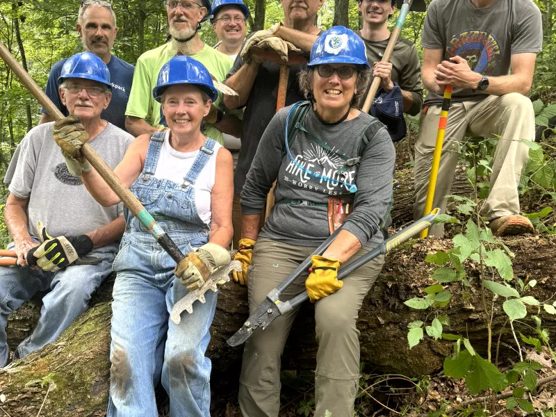 A group of smiling volunteers wearing hard hats and holding tools, posing for a photo on a wooded trail.