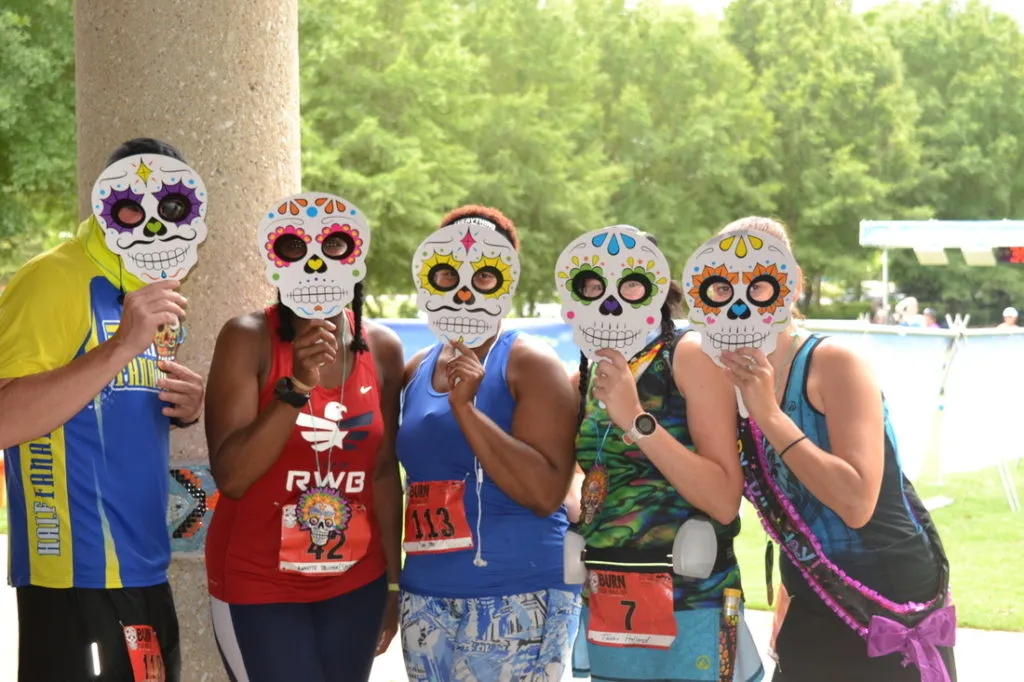A group of five runners wearing colorful Day of the Dead masks at a race event.