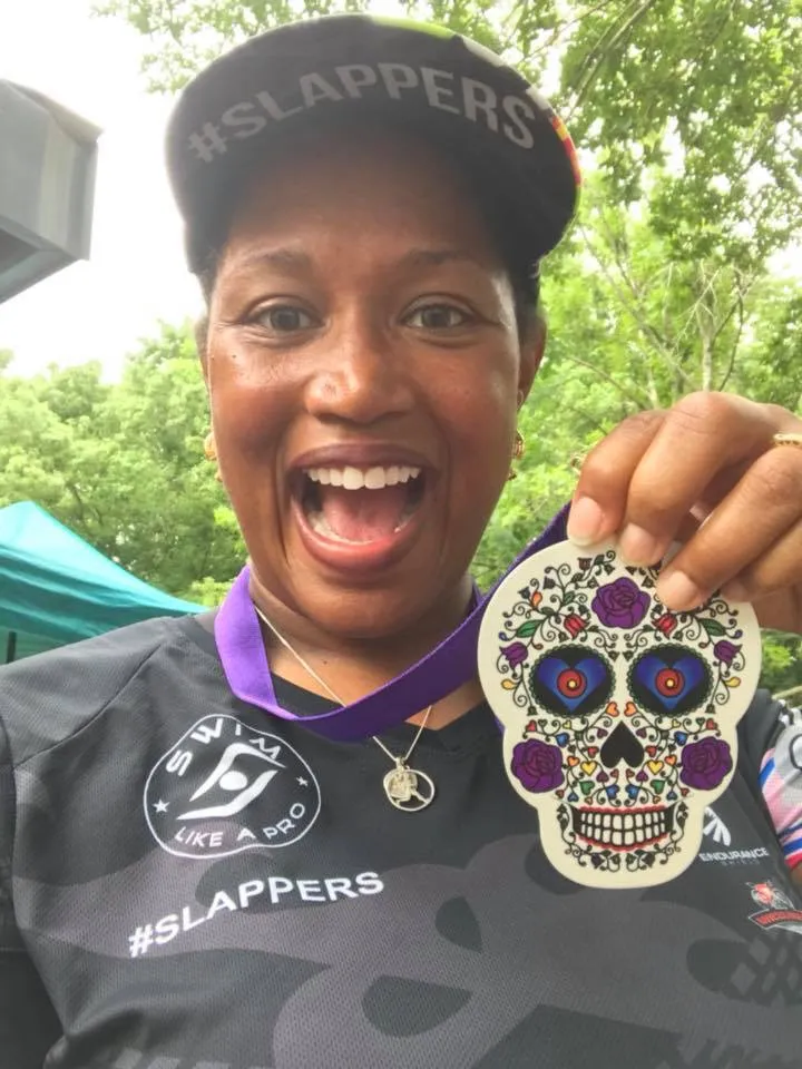 A woman smiles excitedly while holding up a skull-shaped medal after a race.
