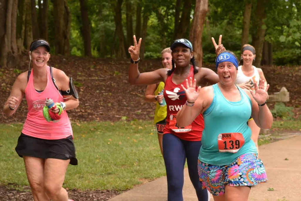 A group of female runners smiling and waving at the camera during a race, with trees and foliage in the background.