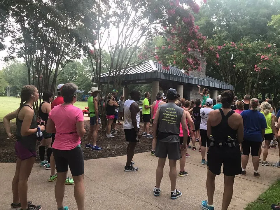 A group of runners in athletic wear gathered outdoors, with trees and a pavilion in the background, appearing to be at the start of a race.