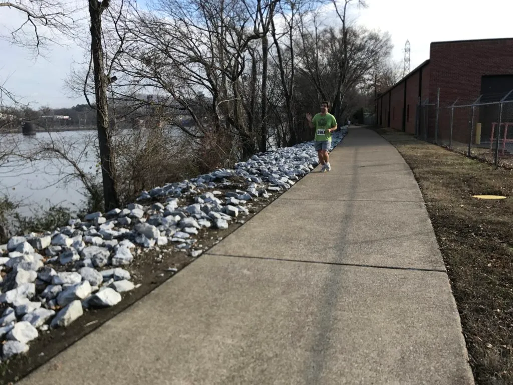 A runner wearing a green shirt and bib number 1983 waves while running on a paved path next to a body of water.