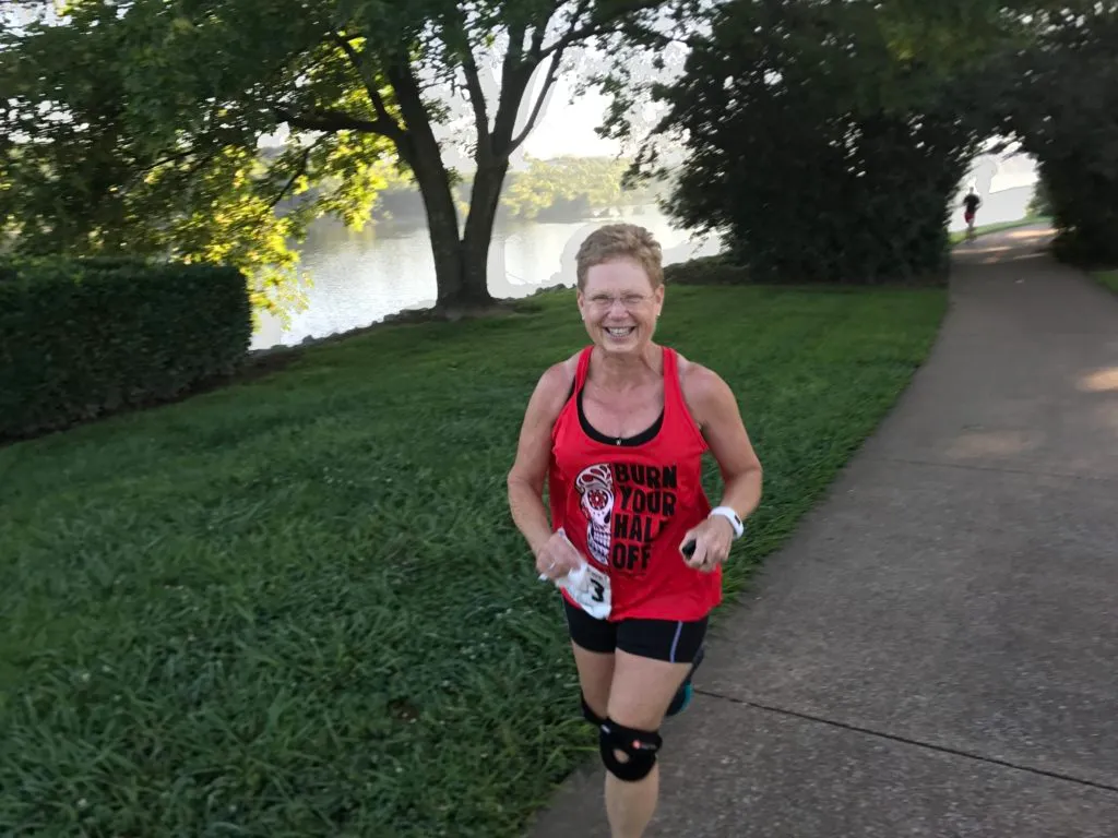 A woman smiles as she runs on a paved path next to a body of water, wearing a red tank top that says "BURN YOUR HALF OFF".
