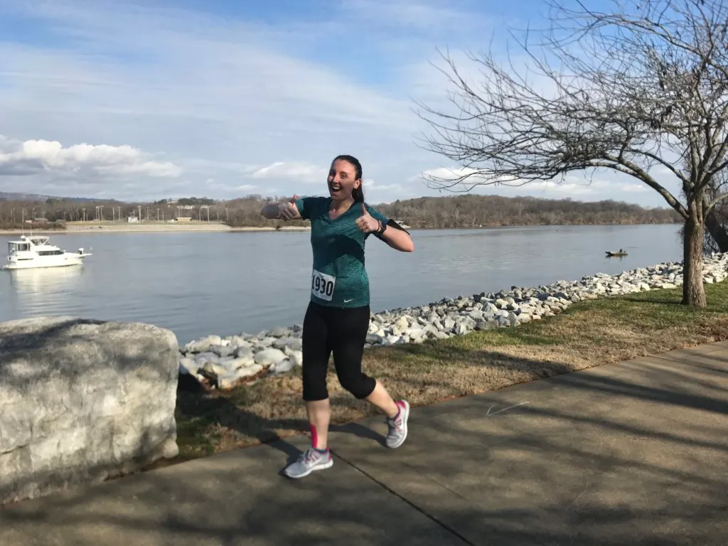 A female runner with bib number 1930 smiles and gives a thumbs up while running along a paved path next to a body of water.