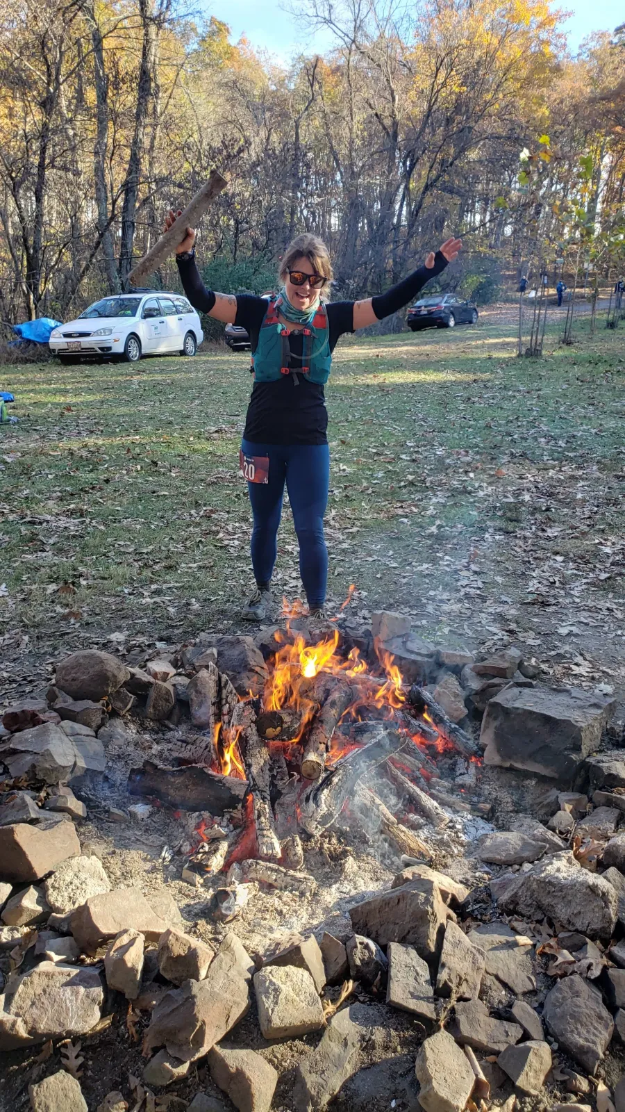 A smiling runner with arms outstretched holds a stick over a campfire, wearing a race bib.