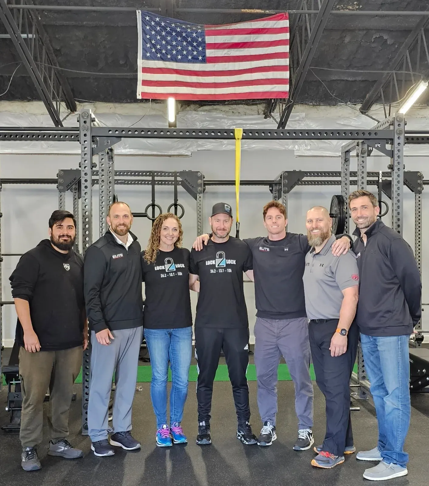 A group of seven people, including men and women, stand together in a gym with an American flag and weightlifting equipment in the background.