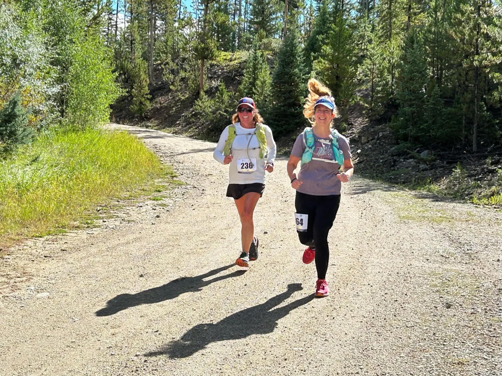 Two female runners smiling as they run on a dirt path surrounded by trees.