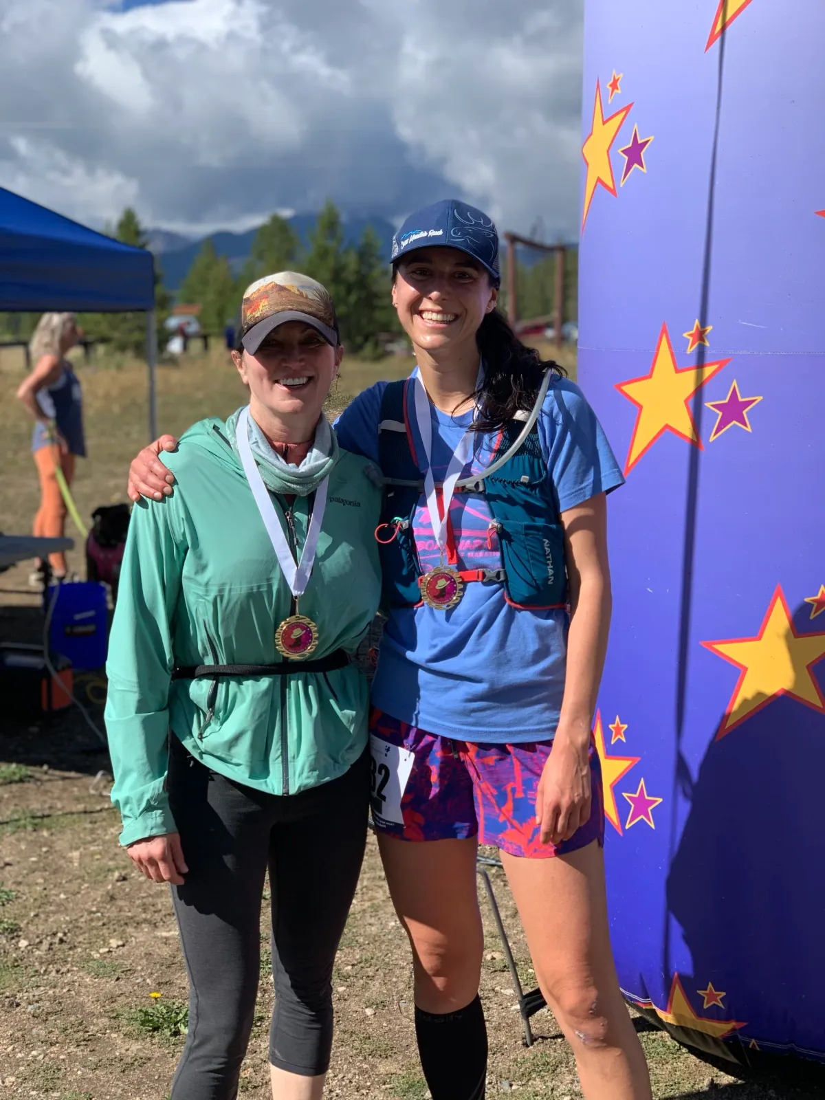 Two smiling women wearing race medals stand next to a colorful inflatable arch decorated with stars.