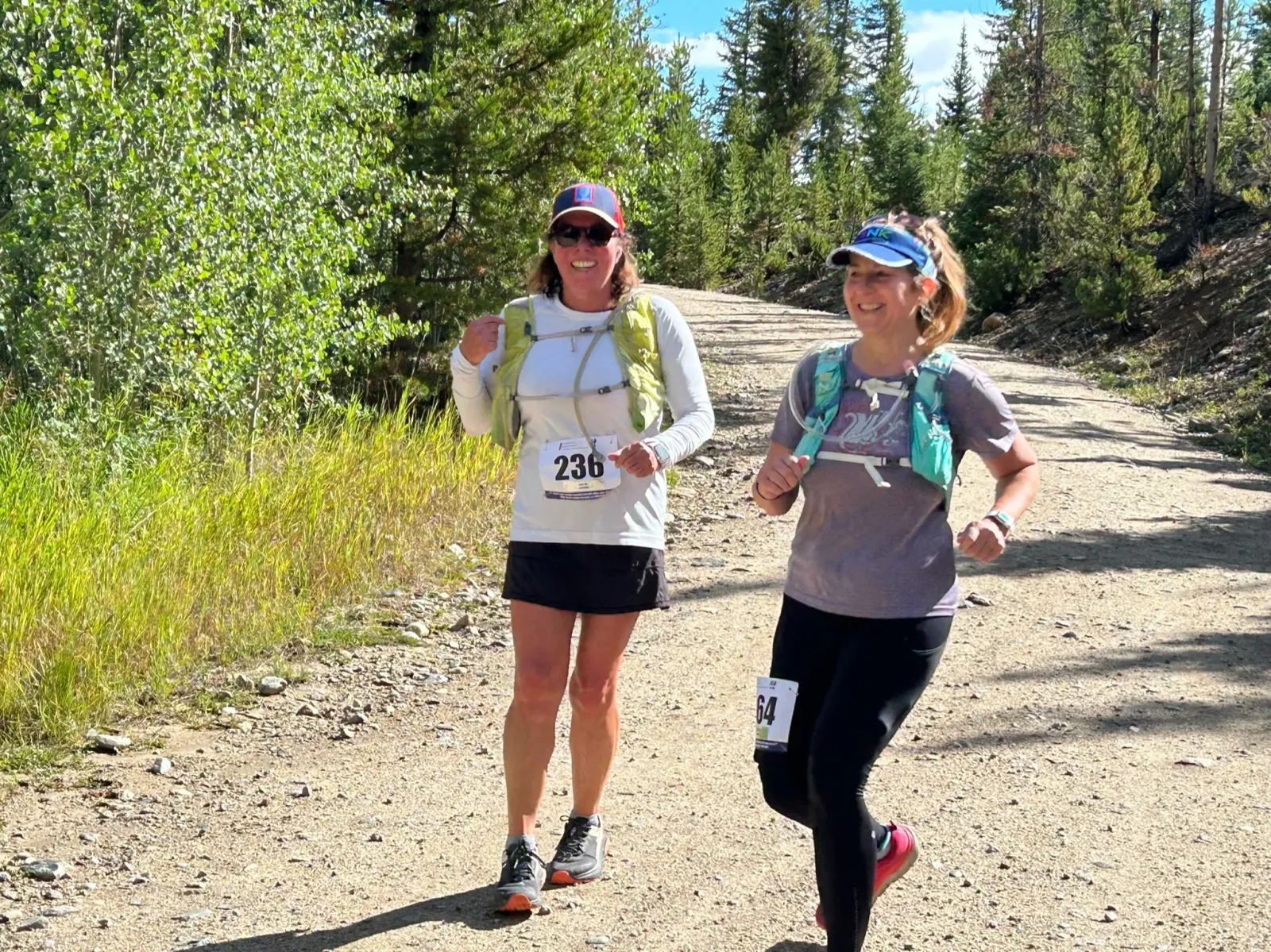 Two women running on a dirt trail surrounded by trees, both smiling and wearing running gear.