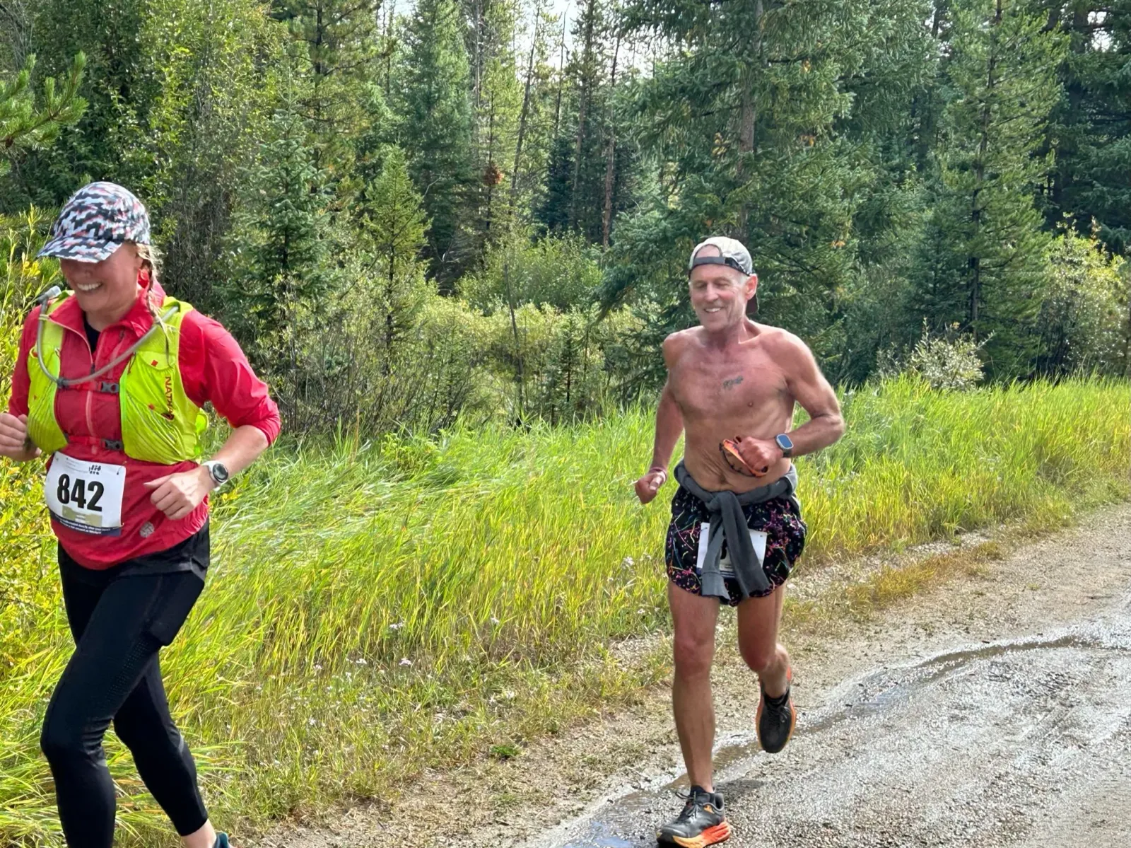 Two runners, one male and one female, are running on a dirt path with trees and tall grass in the background. The female runner on the left is wearing a red shirt, a bright yellow vest, and a patterned cap, with bib number 842 visible. The male runner on the right is shirtless, wearing shorts and a cap, and appears to be smiling.