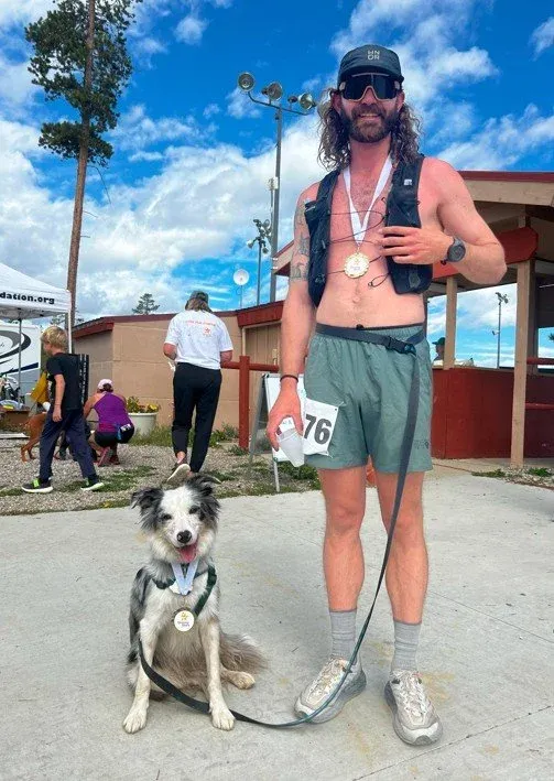 A smiling runner and a dog, both wearing medals, pose for a photo after a race.