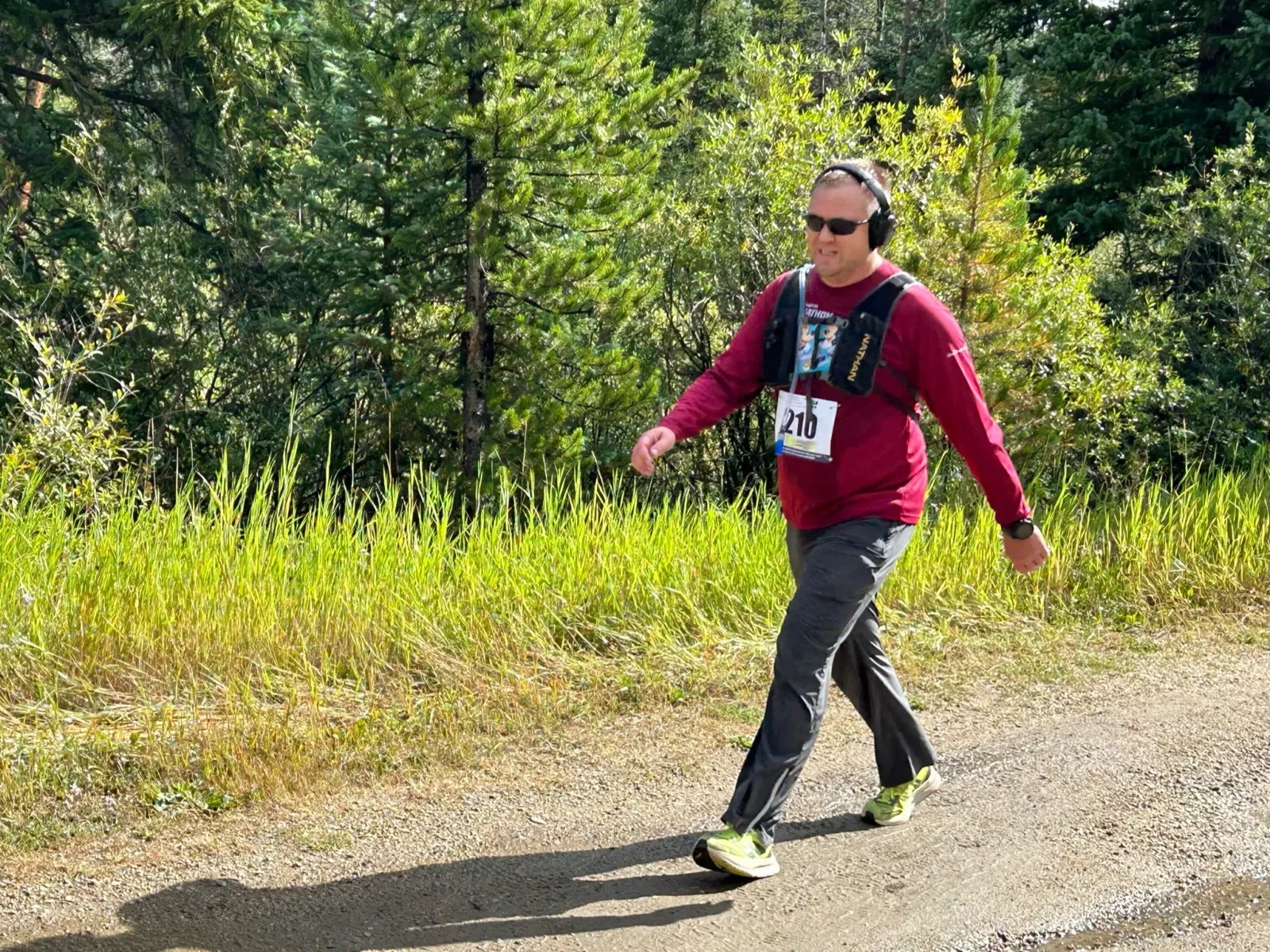 A runner wearing headphones and a bib number walks on a dirt path with trees and tall grass in the background.