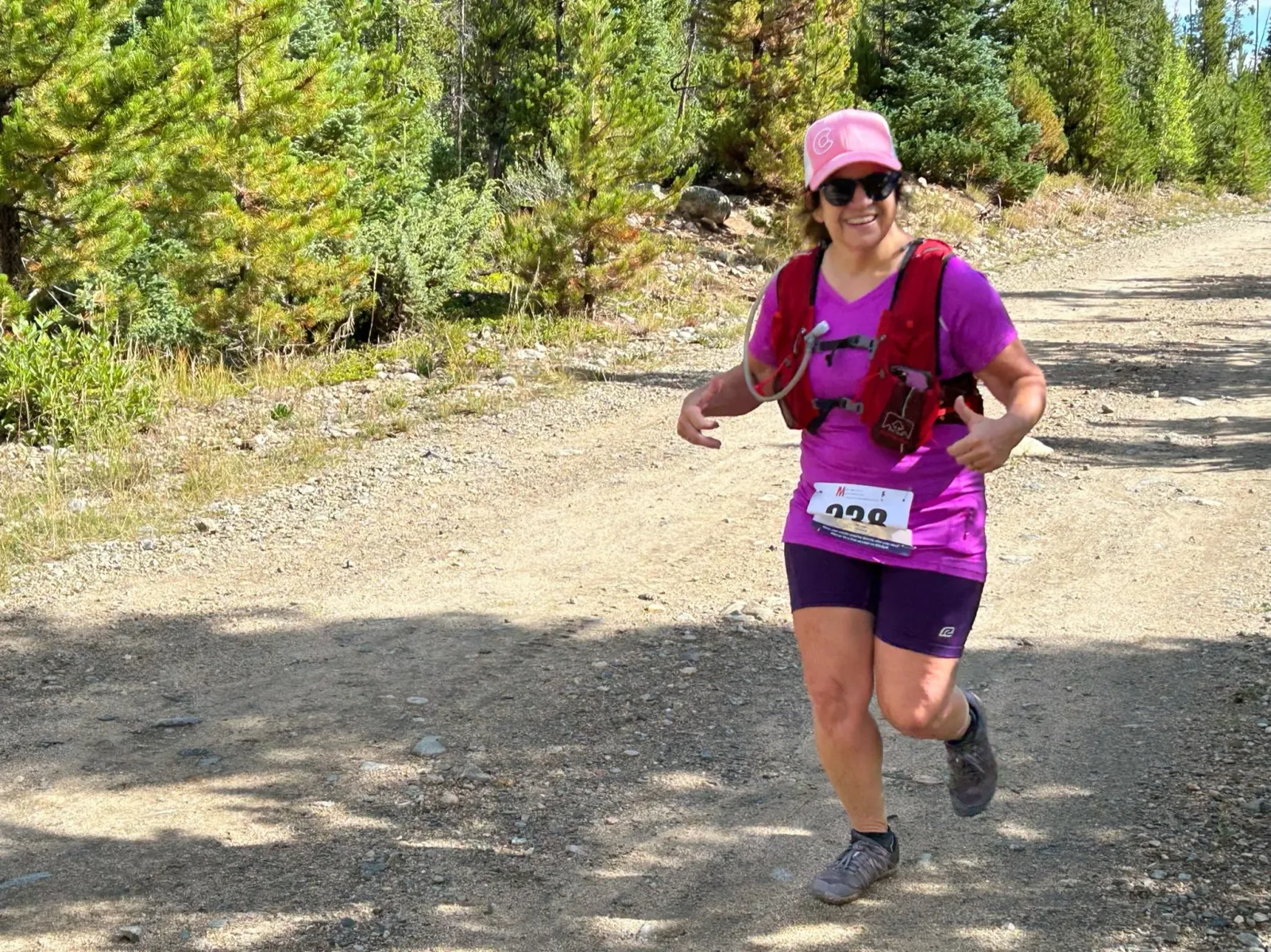 A female runner wearing a pink hat and sunglasses, a purple shirt, and purple shorts, runs on a dirt trail with trees in the background.