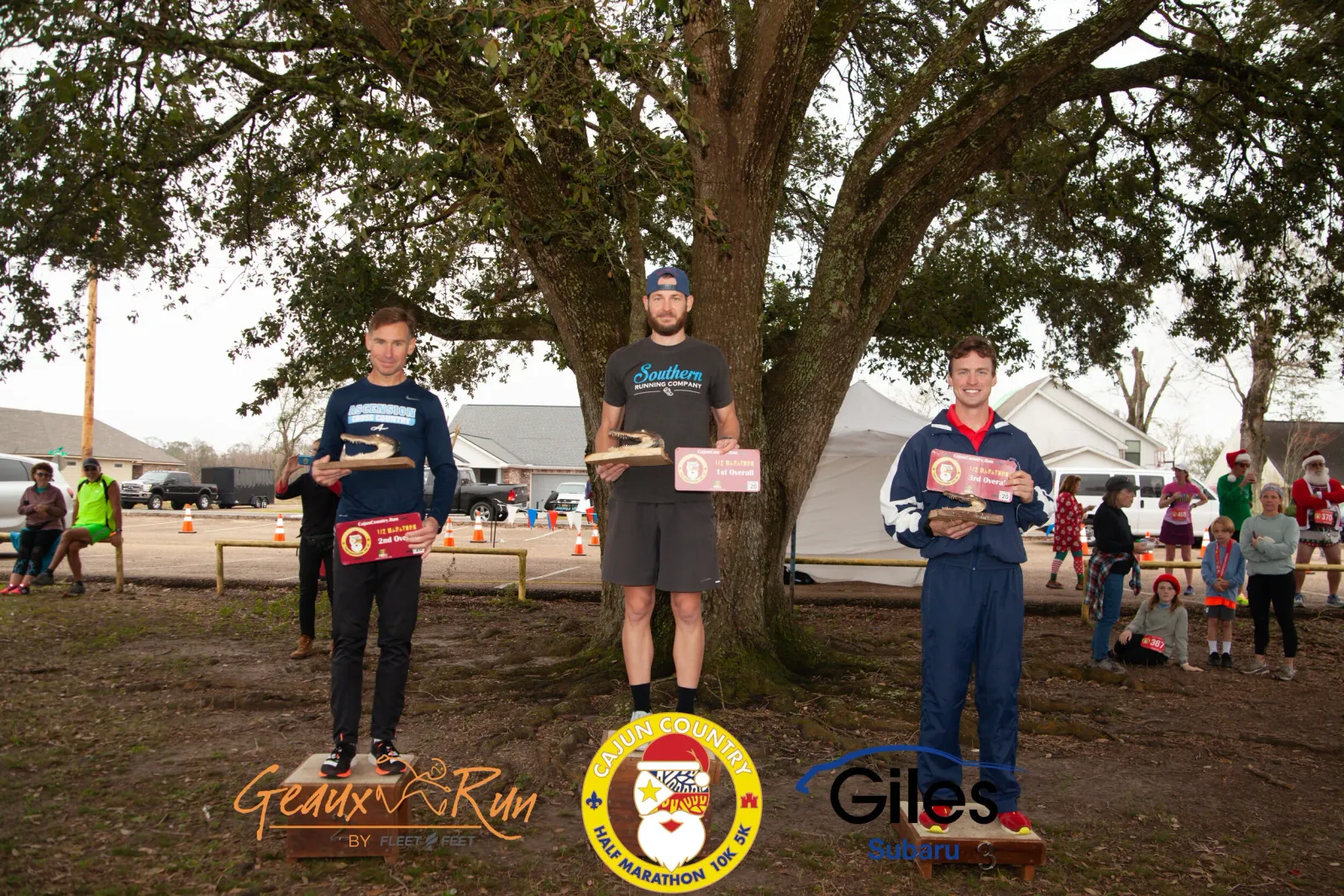 Three male runners on a podium holding awards, with a crowd of people in the background.