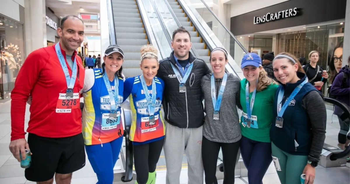 A group of six runners, all smiling and wearing race medals, pose for a photo indoors with escalators in the background.