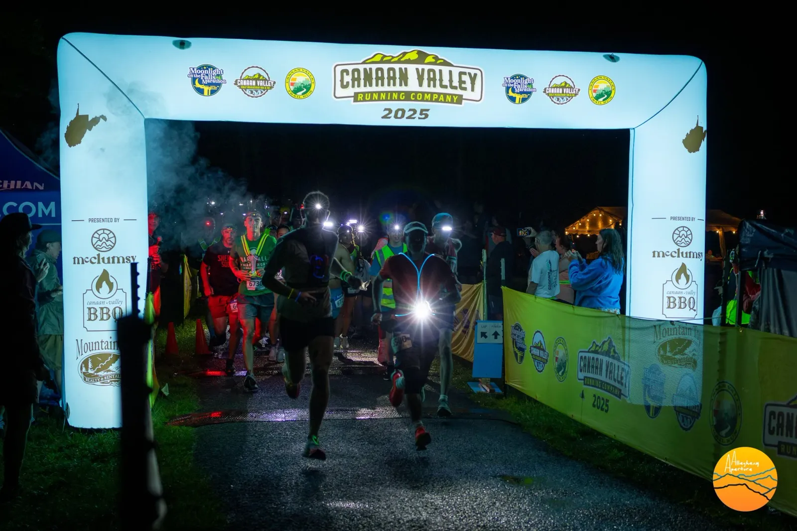 Runners starting a night race under a brightly lit archway with the Canaan Valley Running Company logo.