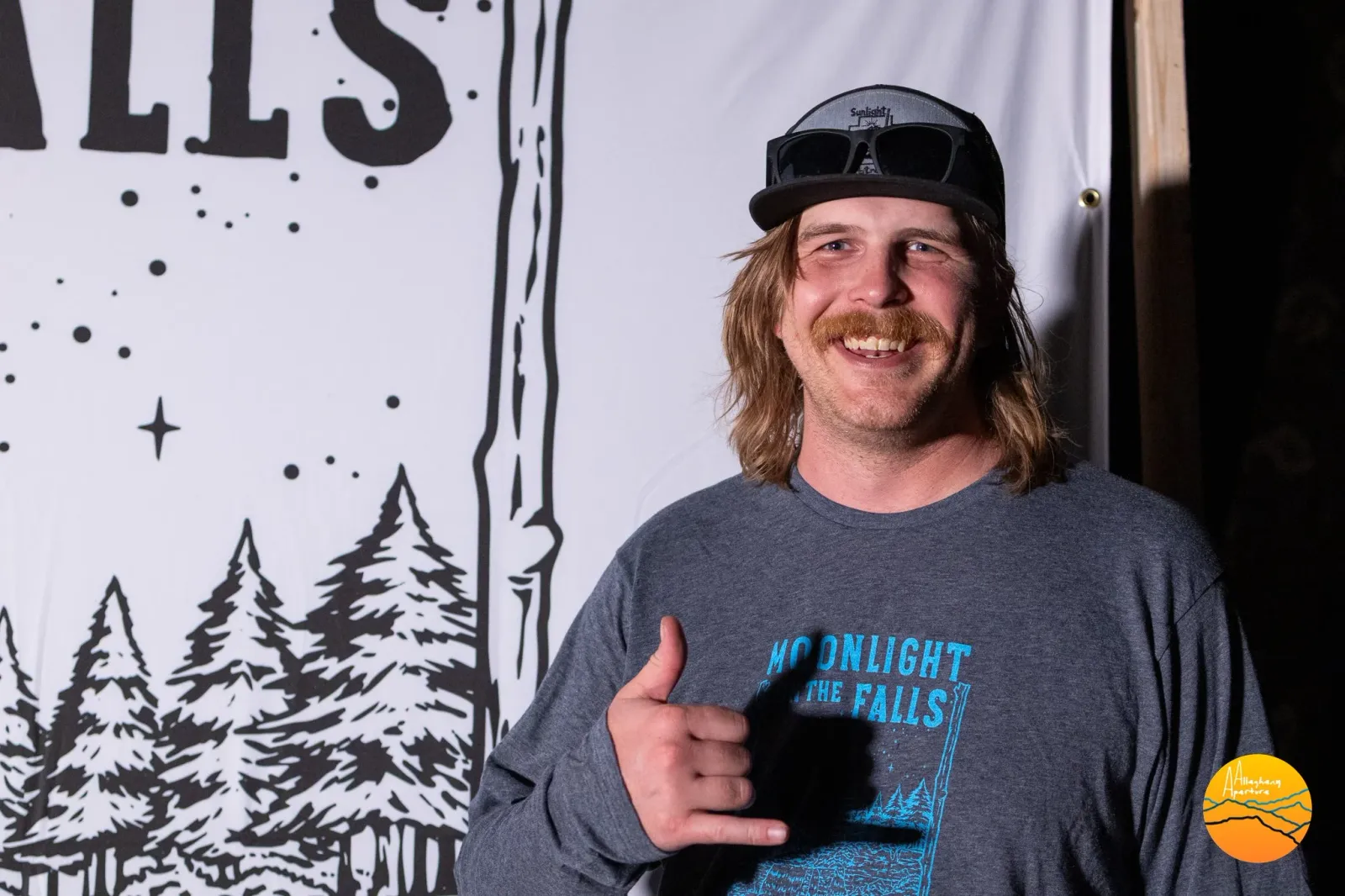 A smiling man with a mustache and a baseball cap gives a shaka sign in front of a banner with trees and text.