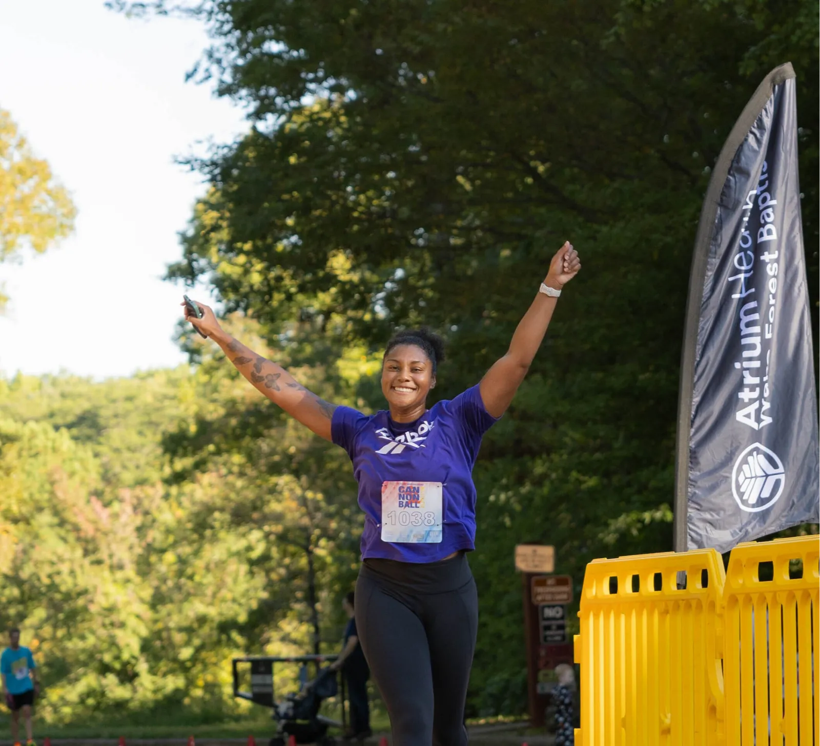 A female runner with her arms raised in celebration, smiling broadly, wearing a purple t-shirt with a race bib number 1038.