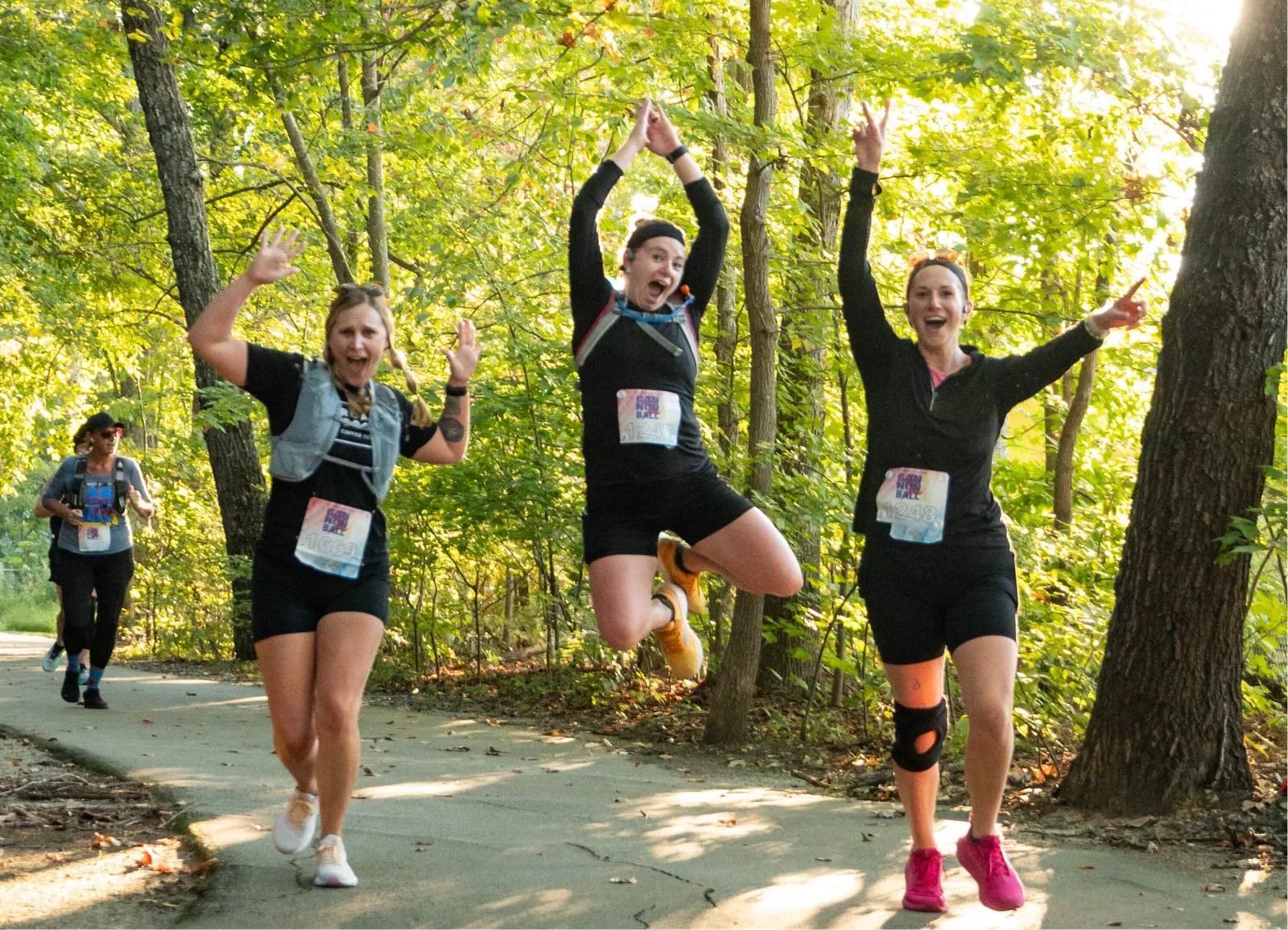 Three women runners joyfully jumping and waving on a paved path surrounded by trees, with sunlight filtering through the leaves.
