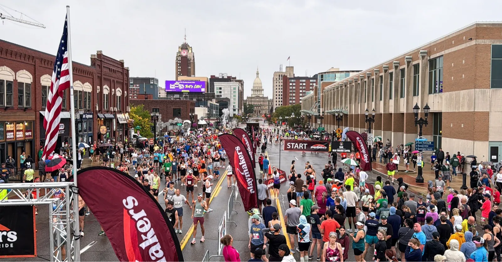A large crowd of runners gathered at the start line of a city race, with buildings and an American flag in the background.