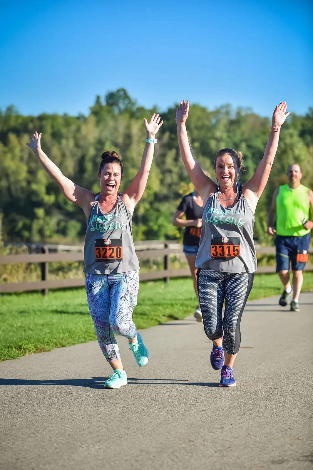 Two women running a race with their arms raised in celebration, wearing bibs with numbers 3220 and 3315.