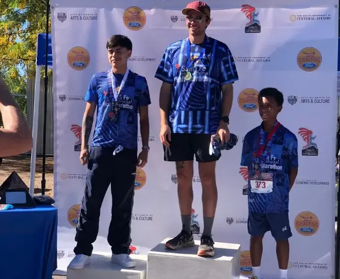 Three runners stand on a podium, wearing medals and race t-shirts, with a backdrop featuring race sponsors and event details.