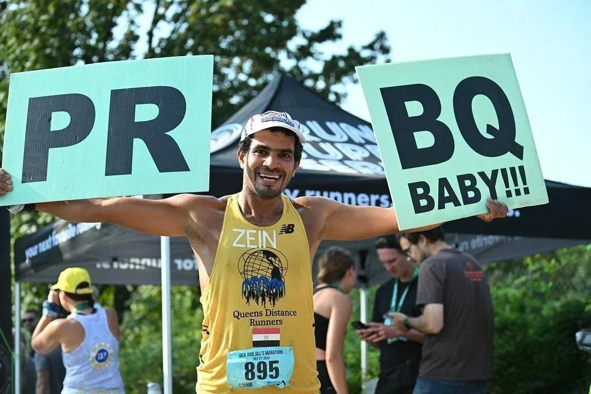 A smiling runner holds up signs that say "PR" and "BQ BABY!!!" at a race.