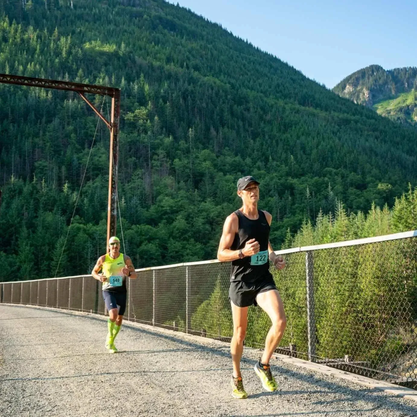Two runners race across a bridge with a lush, forested mountain backdrop.