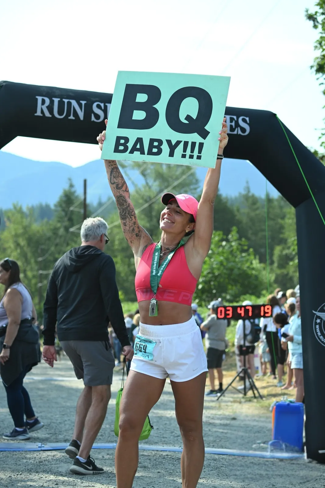 A woman in a pink sports bra and white shorts celebrates at the finish line of a race, holding up a sign that says "BQ BABY!!!" She is wearing a race medal and a bib number.
