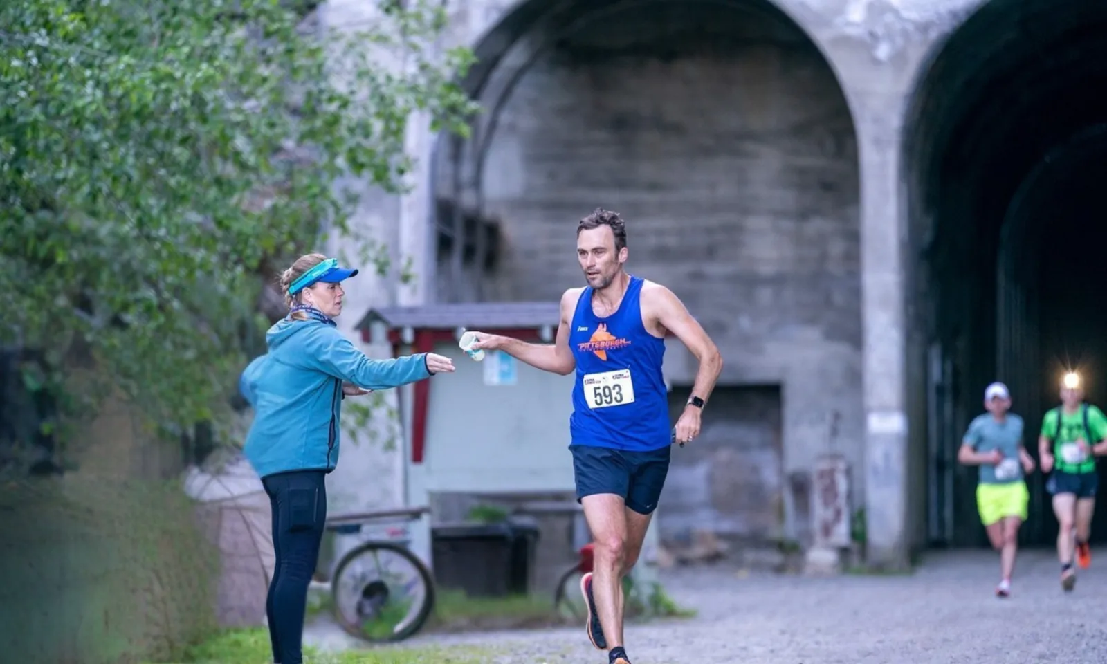 A male runner in a blue tank top with bib number 593 is taking a cup of water from a volunteer during a race, with other runners in the background entering a tunnel.