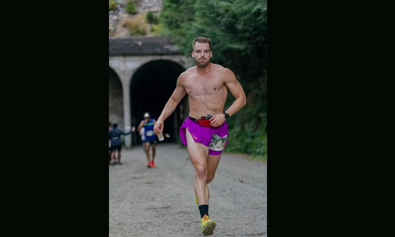 A shirtless male runner with purple shorts runs towards the camera on a dirt path, with a tunnel and trees in the background.