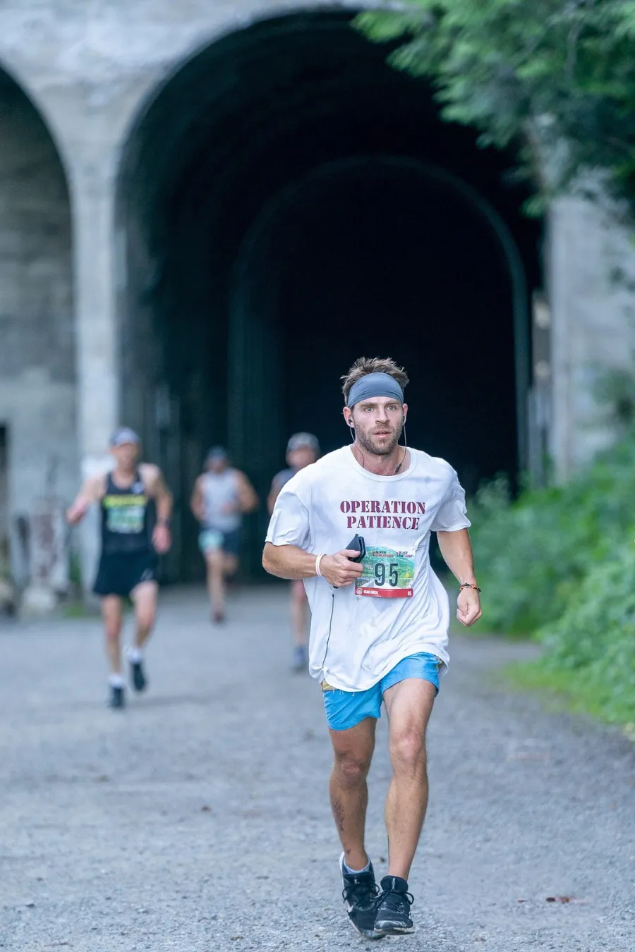 A male runner with a headband and earbuds runs towards the camera, wearing a white t-shirt with 'OPERATION PATIENCE' and bib number 95, with other runners and a tunnel in the background.