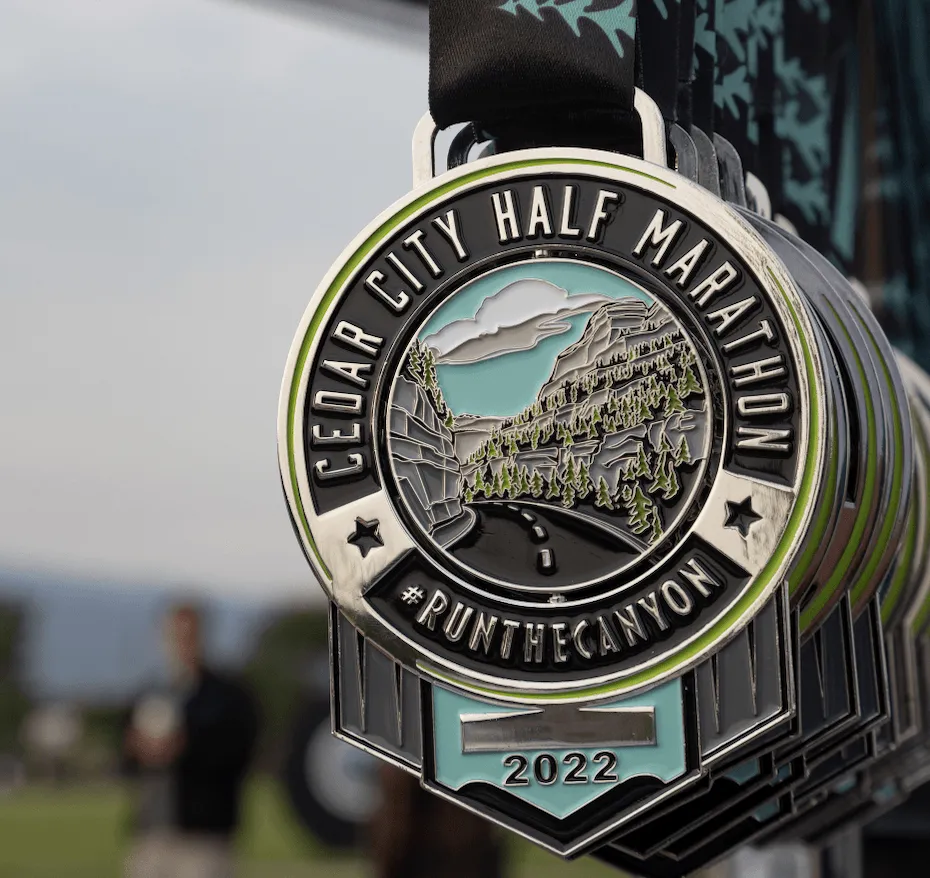 A close-up of the Cedar City Half Marathon medal, with a scenic canyon road design, hanging with other medals. Blurred runners are visible in the background.