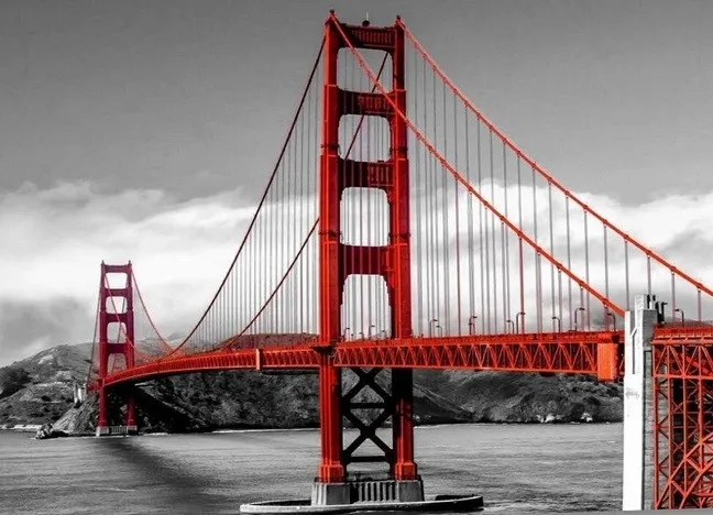 A striking red suspension bridge against a grayscale background, with water in the foreground and hills in the distance.