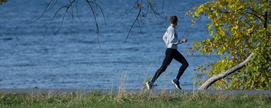 A runner runs along a path next to a body of water, with trees and foliage in the foreground and background.