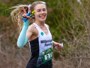 A female runner with blonde hair tied in a ponytail smiles at the camera while running, holding up a peace sign with her left hand and a water bottle in her right.
